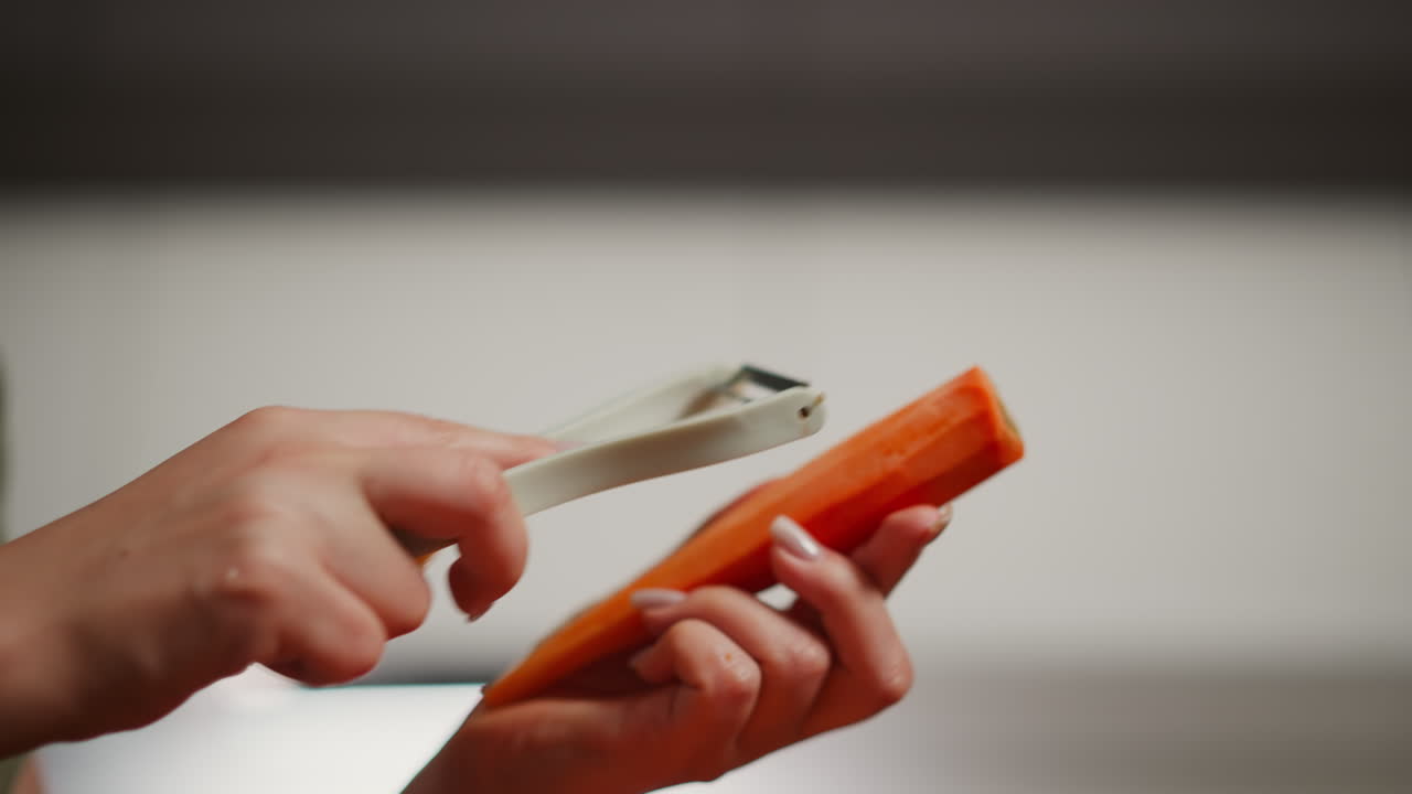 Close up of lady hand with polished nail using white peeler to shave orange carrot, blur background suggests modern kitchen, focus on precise cutting action in clean cooking environment