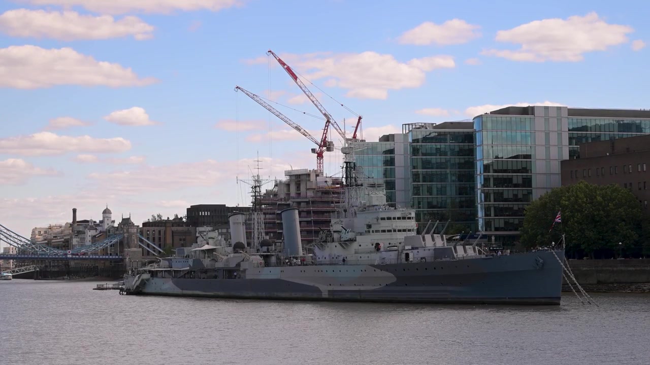 Historisch oorlogsschip HMS Belfast ligt afgemeerd aan de Theems, terwijl bouwkrane oprijzen tussen nabijgelegen glazen kantoren onder een heldere hemel.