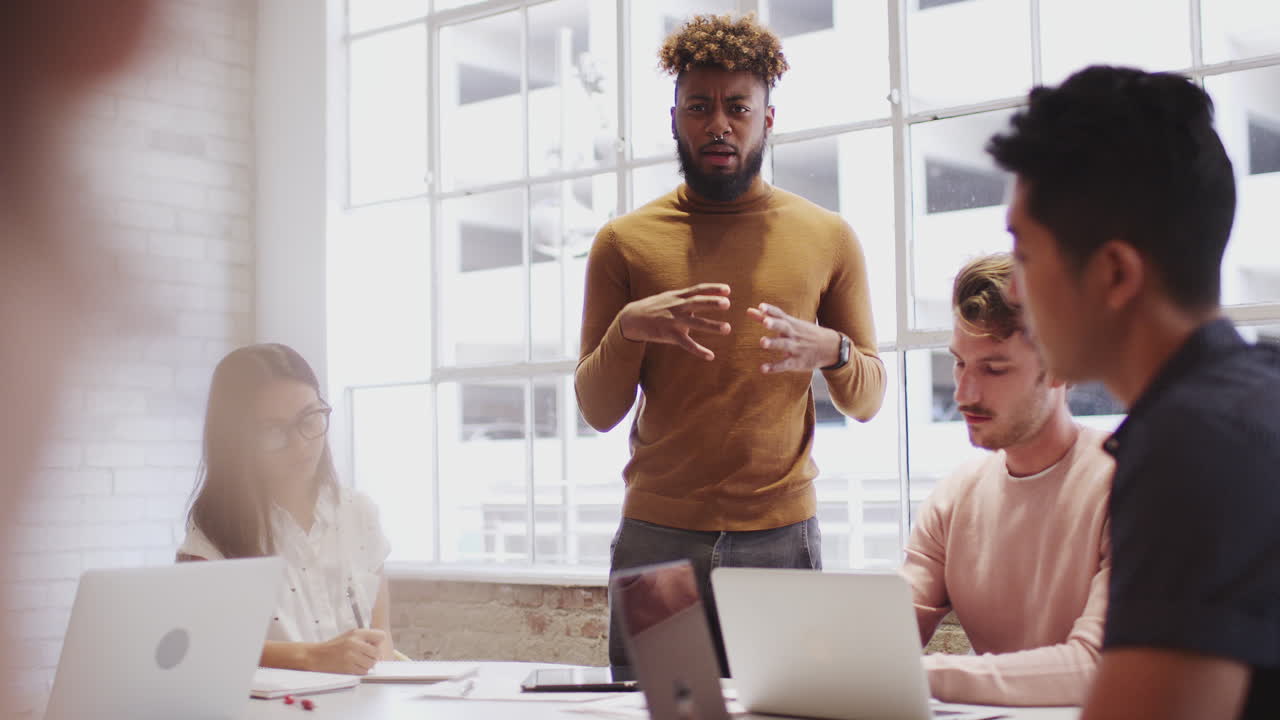 Black male creative stands addressing colleagues at a table in a meeting room, selective focus