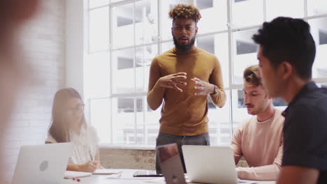 Black-male-creative-stands-addressing-colleagues-at-a-table-in-a-meeting-room,-selective-focus