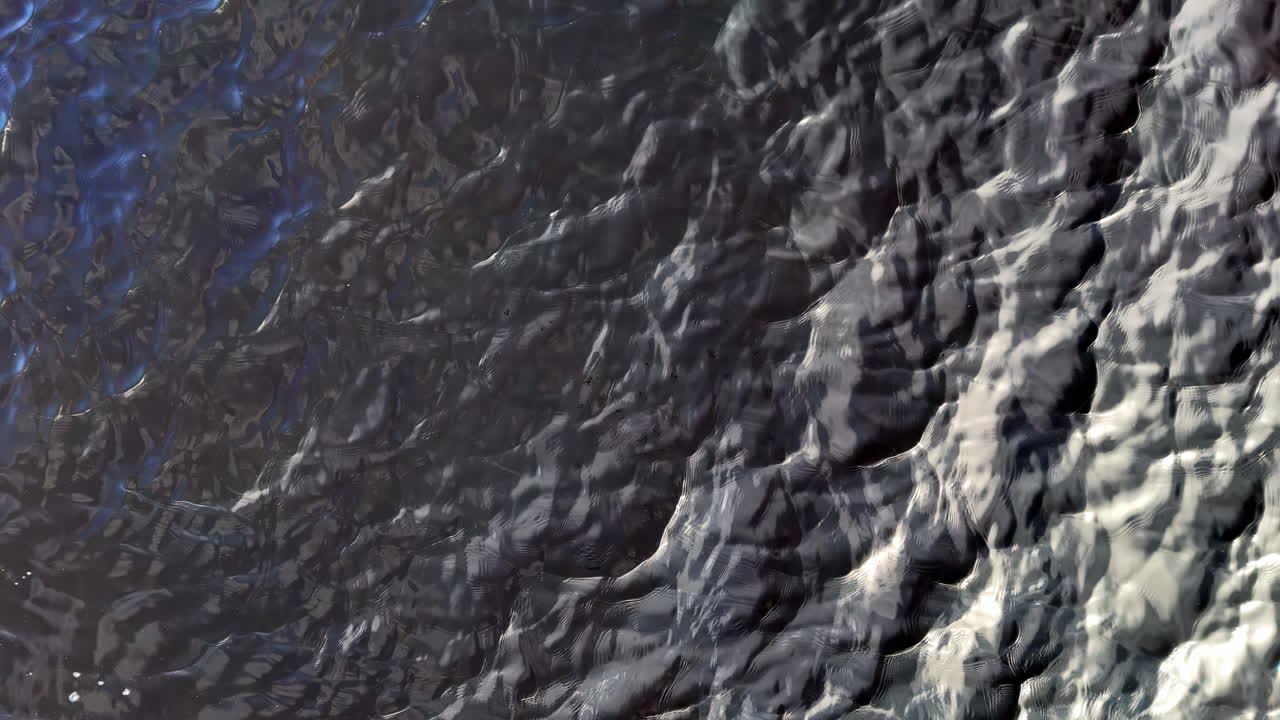 A textured floodwater surface catches sharp light and dark contrasts, creating a jagged ripple effect. This abstract aerial view captures nature’s chaotic force and visual complexity