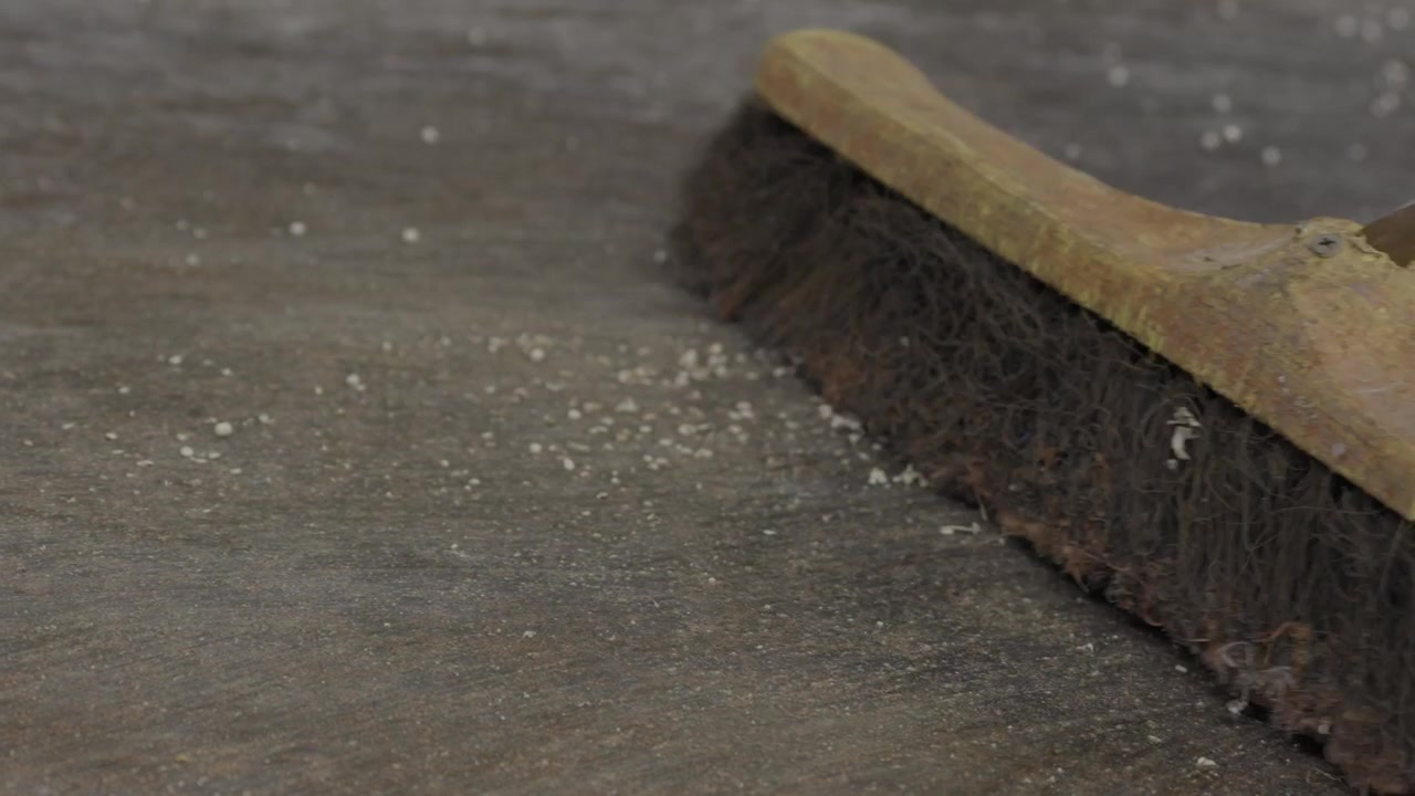 Trabalhador varre grânulos de plástico branco reciclado espalhados pelo chão cinzento da fábrica com vassoura em close-up