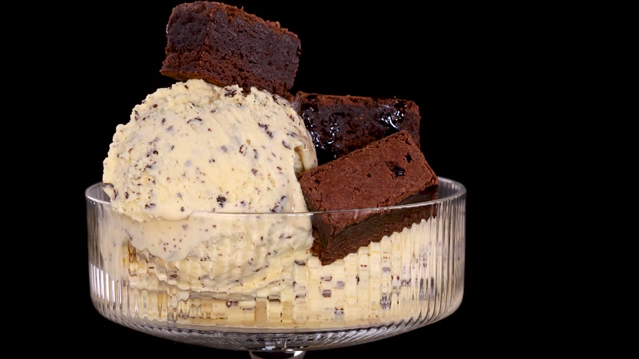 A hand places chocolate brownie pieces onto a scoop of vanilla ice cream with chocolate chips in a glass bowl, against a black background with bright, even lighting