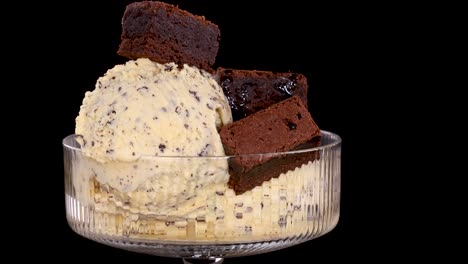 a hand places chocolate brownie pieces onto a scoop of vanilla ice cream with chocolate chips in a glass bowl, against a black background with bright, even lighting