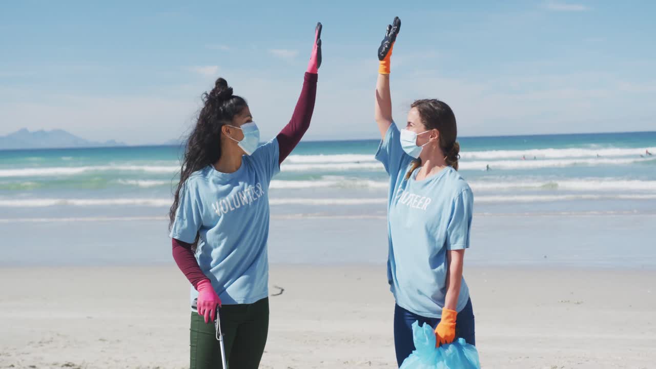 두 명의 다양한 여성이 자원봉사자 티셔츠와 얼굴 마스크를 입고 해변에서 쓰레기를 모으고 있습니다.