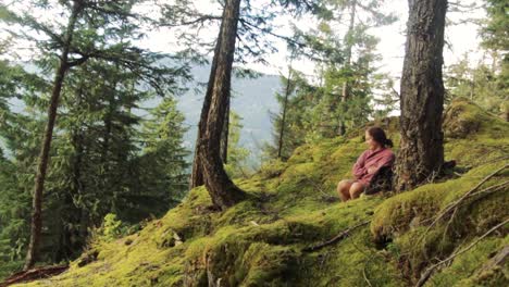 A-beautiful-young-adult-female-is-sitting-in-the-mossy-forest-near-a-tree-and-enjoying-sunshine,-nature-and-calmness