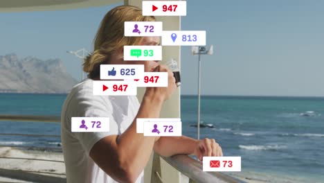 animation of social media icons and numbers flying over caucasian man drinking coffee by seaside