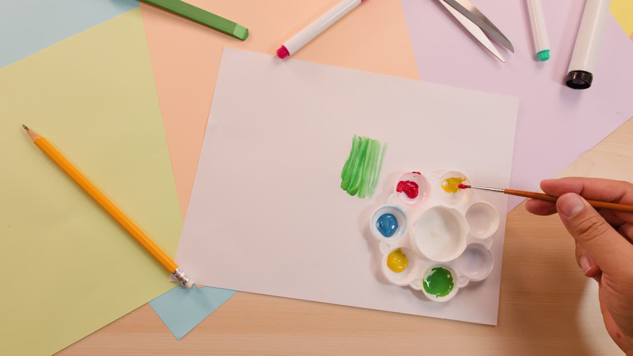 Hands mix red and yellow paint on palette above blank paper, under soft even lighting