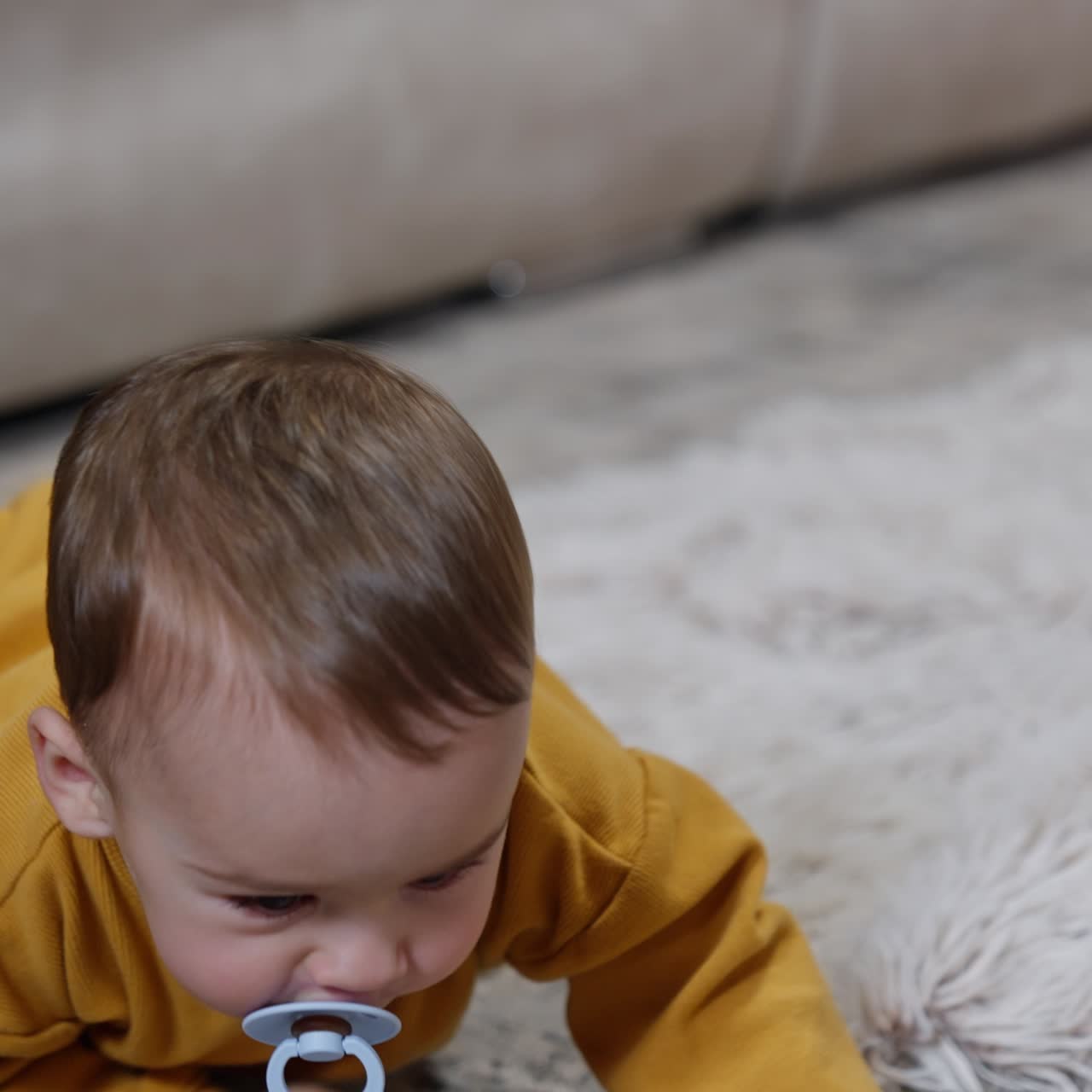 Baby boy lies on the floor holding legs on the sofa. Little kid with pacifier stands on all four and crawls away