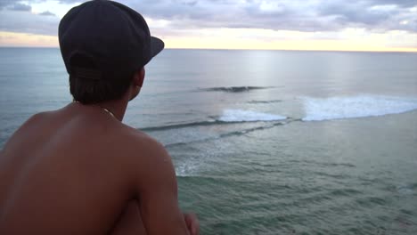 slow motion man sits on edge of cliff looking out at waves crashing in ocean