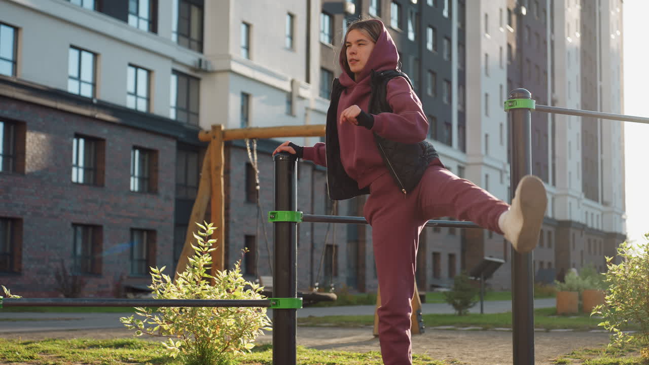 Strength trainer in hoodie grips metal pull up bar while raising side leg high during outdoor workout in fitness park under bright sun autumn foliage modern backdrop showcasing flexibility resilience