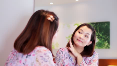 beautiful asian woman looks into bathroom mirror touches her lush black hair, enjoys her looks