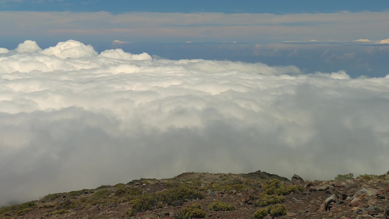 Sweeping footage of Haleakala National Park summit, Maui, shows a rocky volcanic ridge overlooking a dramatic sea of clouds under vibrant daylight in a wide angle landscape view.