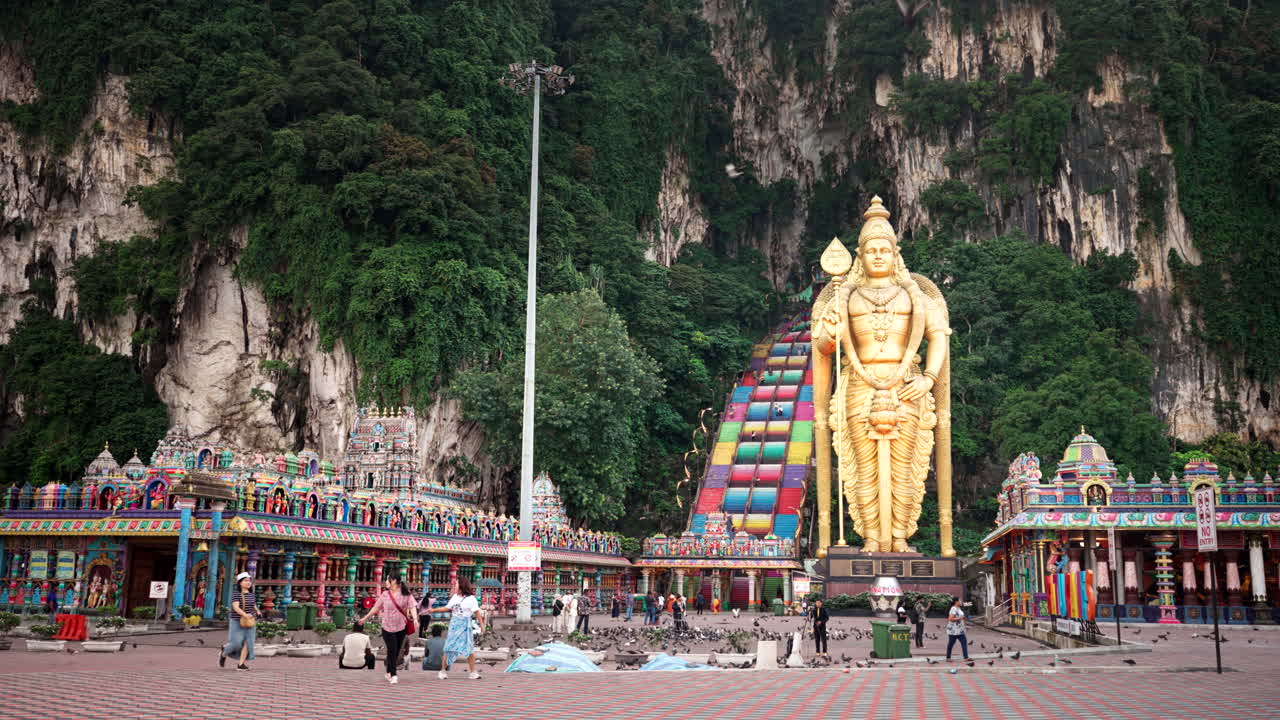 People Feeding Pigeons On Ground Of Batu Caves With Murugan Sculpture At Entrance. Gombak, Selangor, Malaysia. wide shot
