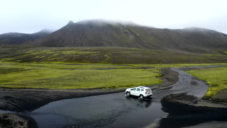 Imágenes-Aéreas-De-Drones-De-Un-Camión-4x4-Blanco-Conduciendo-A-Través-De-Un-Río-Que-Cruza-Las-Tierras-Altas-De-Islandia