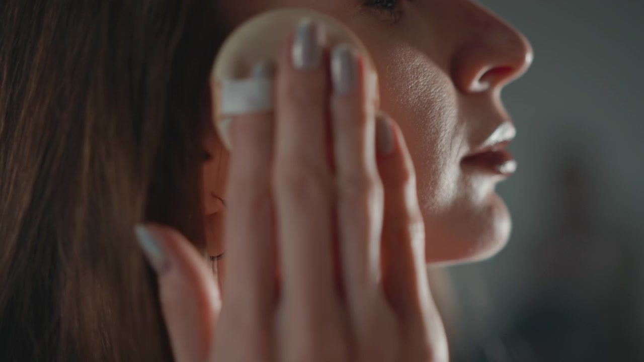 Close profile view shows young woman wiping cheek and lower eye area with round cotton pad in slow circular strokes, soft side light revealing skin texture, focused grooming and cleansing routine