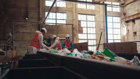 Side-view-of-waste-recycling-plant-workers-in-special-uniforms-laying-out-and-sorting-plastic-bottles-based-on-their-color-while-standing-near-a-moving-conveyor-belt-at-a-big-old-plant