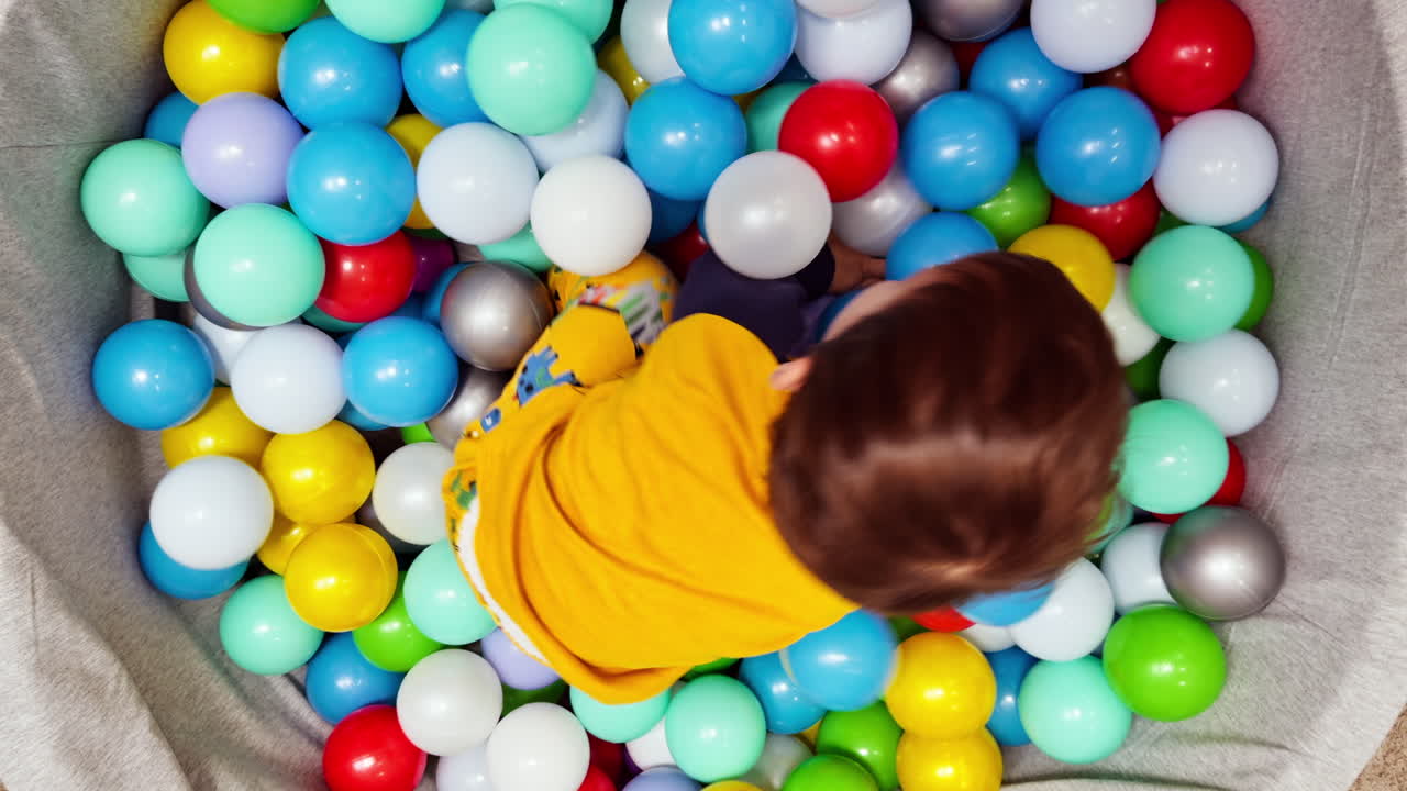 Happy playful kid lies in the pool among the colorful balls. Top view on the child playing in dry basin.