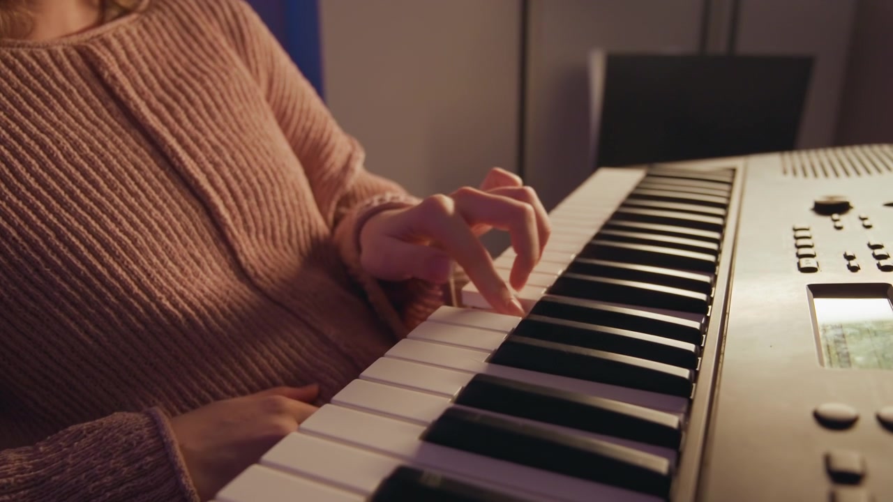 Close-up shows both hands playing electronic keyboard with curved fingers, visible key presses, and warm side light in cozy indoor setting, suggesting calm home music practice and gentle rhythm