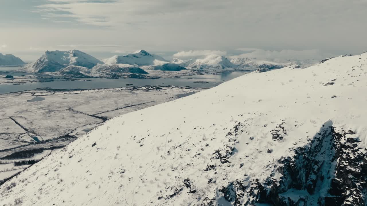 A Snowy Mountain Slope Offers a Stunning View of the Fjords and Peaks in the Lofoten Islands, Norway - Aerial Pullback Shot