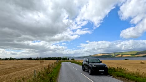 a black vehicle travels along a narrow country road bordered by hayfields and rolling hills under partly cloudy skies, with steady camera framing and natural daylight