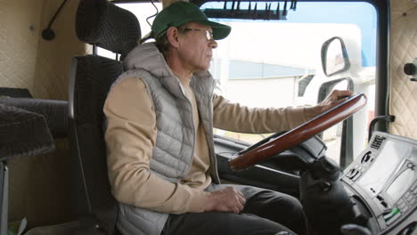 side view of older worker wearing cap and vest driving a truck in a logistics park 1