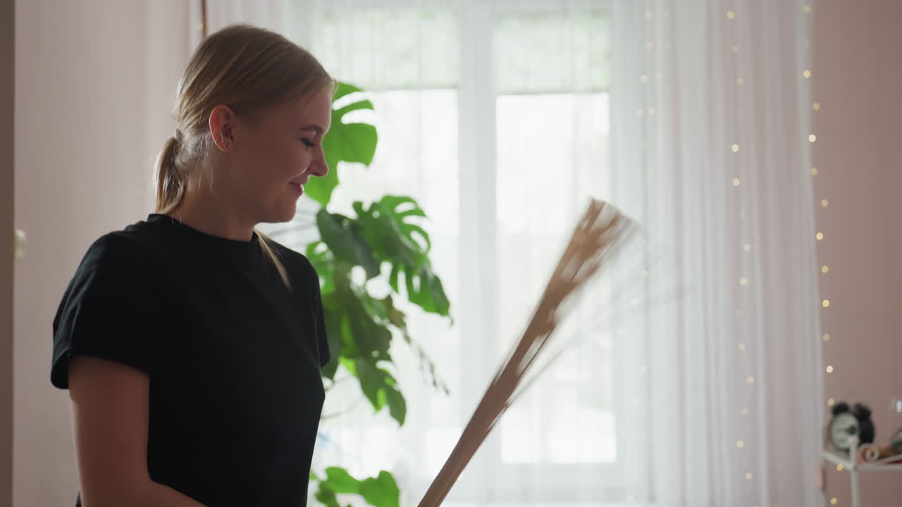Massage therapist with blond hair smiles gently while using bamboo broom on client in softly lit spa setting with green foliage and sheer curtains glowing in daylight atmosphere
