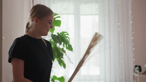 massage therapist with blond hair smiles gently while using bamboo broom on client in softly lit spa setting with green foliage and sheer curtains glowing in daylight atmosphere