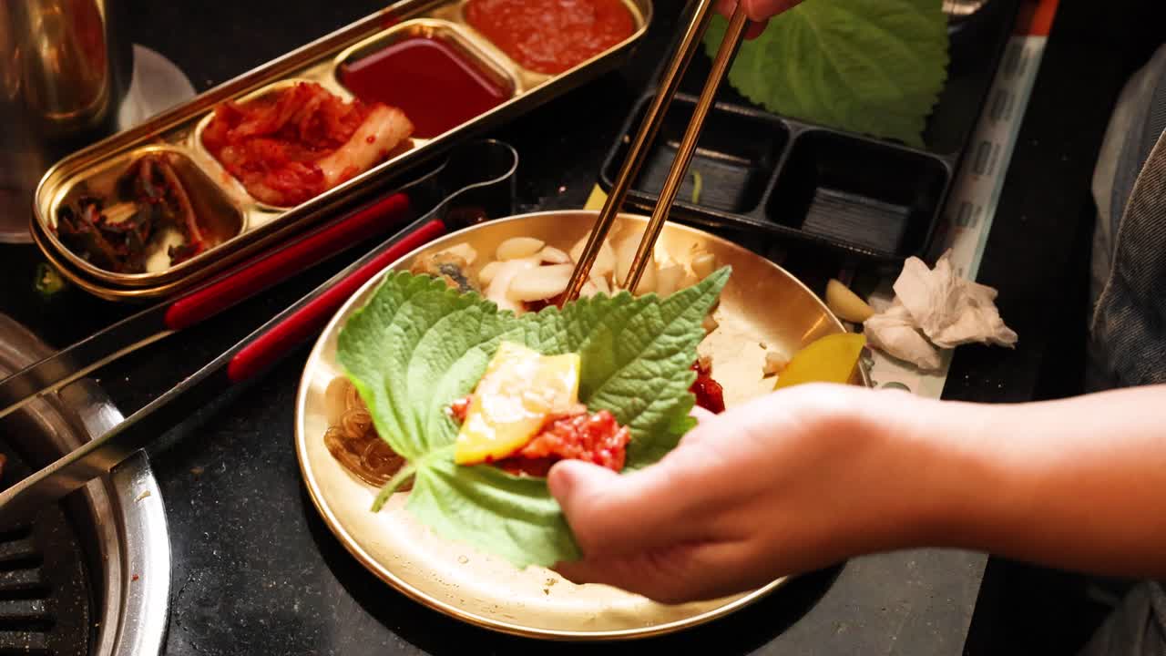 Hand prepares Korean BBQ wrap with perilla leaf, radish, garlic, and beef under warm lighting