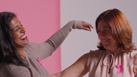 retrato de estudio de dos mujeres maduras que llevan cintas rosadas de concienciación sobre el cáncer de mama abrazándose contra un fondo rosa