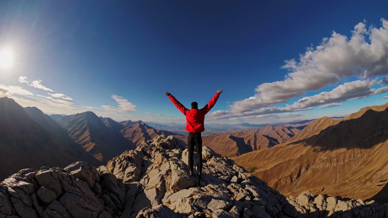 Toma de video aérea de una persona con chaqueta roja de pie en la cima de una montaña, capturando una vasta