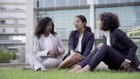 joyful female employees sitting on green grass and talking