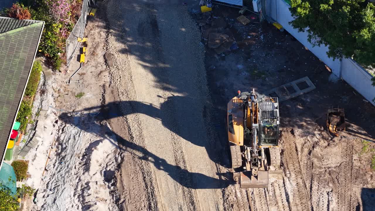 Aerial view of excavator leveling dirt road in bright daylight, steady overhead camera, construction zone