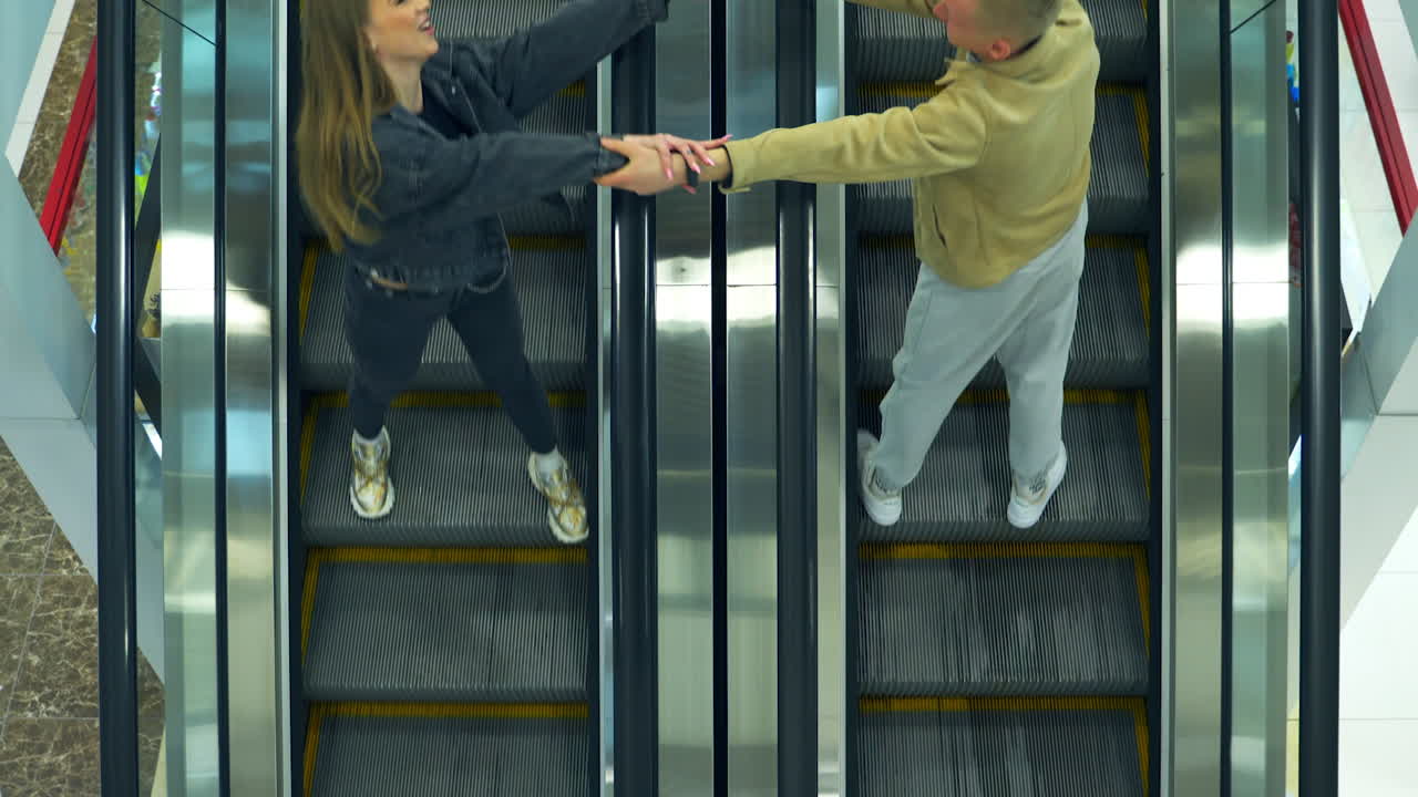 Modern escalator stairs moving quickly in the mall. Woman and man going by opposite lines stretch their hands to each other. Top view.