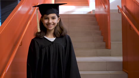 Retrato-De-Una-Feliz-Estudiante-De-Preescolar-Con-Toga-Y-Birrete-Bajando-Las-Escaleras,-Sosteniendo-Un-Diploma-De-Graduación-Y-Mirando-La-Cámara-1