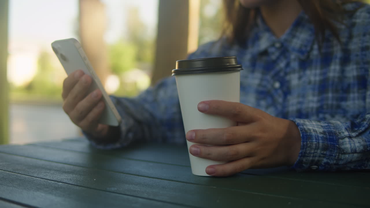 Woman is chatting with friends with phone in the park. Student girl sitting outside drinking coffee and communicating wit smartphone.