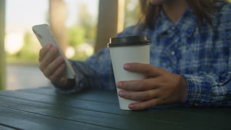 woman is chatting with friends with phone in the park. student girl sitting outside drinking coffee and communicating wit smartphone.
