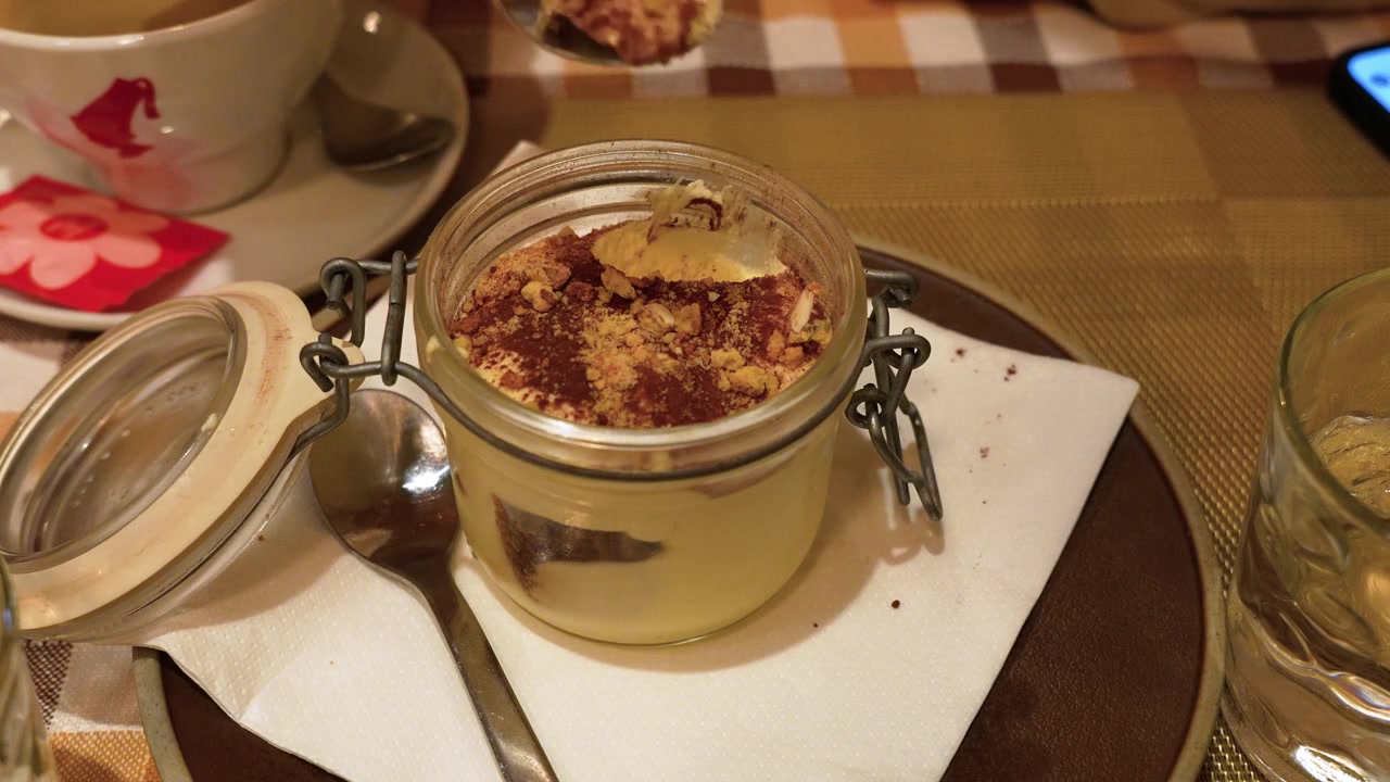 Warm restaurant lighting highlights creamy tiramisu in a glass jar topped with cocoa and nuts. The close-up shot captures spoonfuls being served, evoking inviting dessert moments. (199)