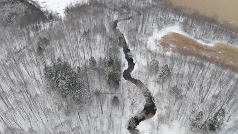 stream winding through snowy wooded landscape between brown farm fields in winter