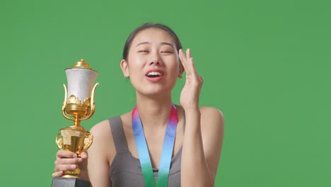 close up of asian woman with a gold medal holding a gold trophy and yelling positively with hand over mouth on green screen background in the studio