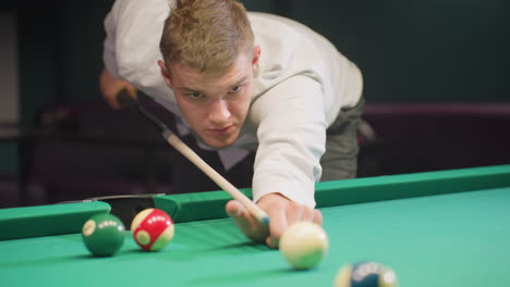 close-up front view of game man in white shirt with serious face, leaning over green pool table in dimly lit room. gripping cue stick with focus, ready to strike white cue ball