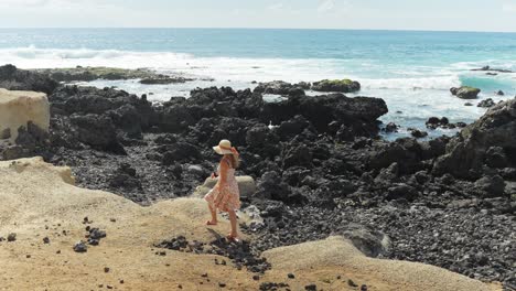 Woman-walking-over-sand-and-black-rocks-near-the-seashore-while-wearing-floral-dress,-static-slow-motion