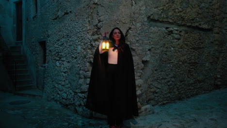 mujer misteriosa con linterna y capa por la noche en una calle de la ciudad medieval