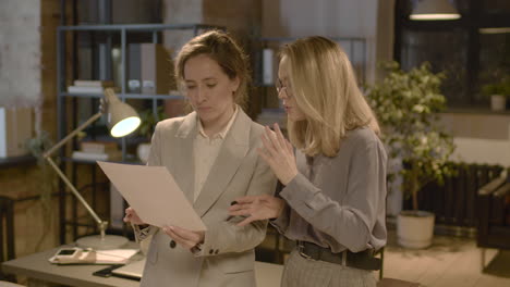 Two-Female-Coworkers-Talking-And-Reading-A-Document-Standing-In-The-Office