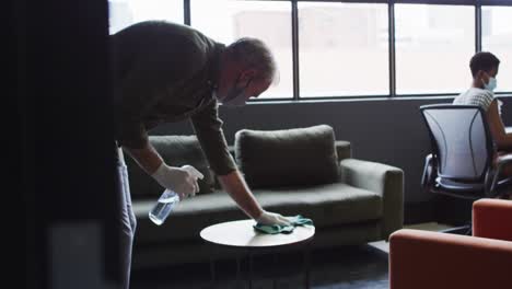 Senior-caucasian-man-wearing-face-mask-amd-gloves-sanitizing-table-in-office