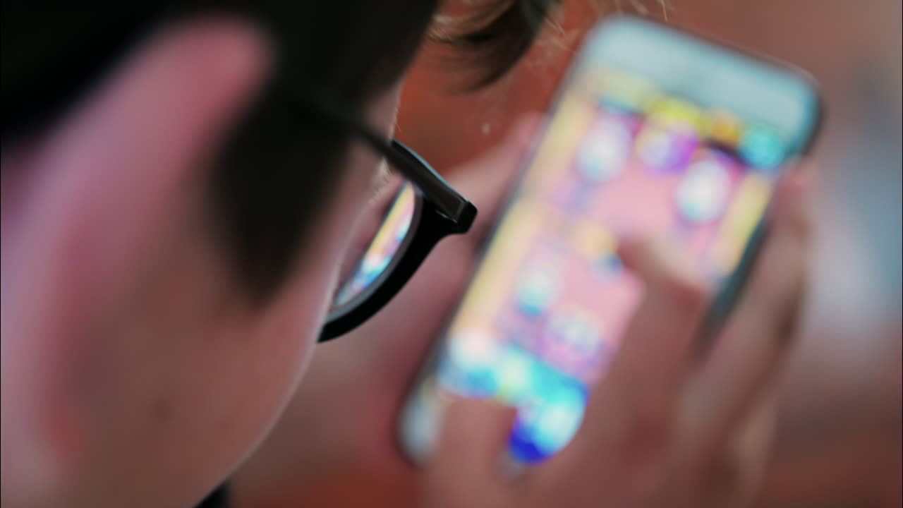 Over-the-shoulder close-up of a kid with glasses playing on a smartphone indoors