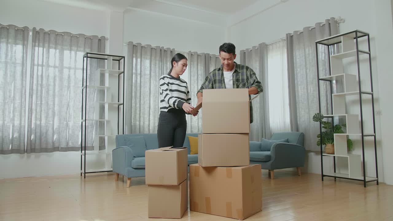 젊은 아시아 커플이 새로운 집으로 이사하고 카드보드 상자를 열고 그 안에 있는 물건들을 꺼내고 있습니다.