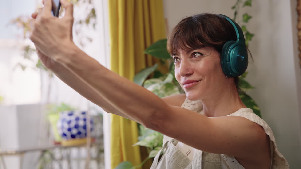 Donna che si scatta un selfie con lo smartphone mentre ascolta musica con le cuffie a casa. Stile di vita quotidiano, social media e interazione digitale in un ambiente interno accogliente.