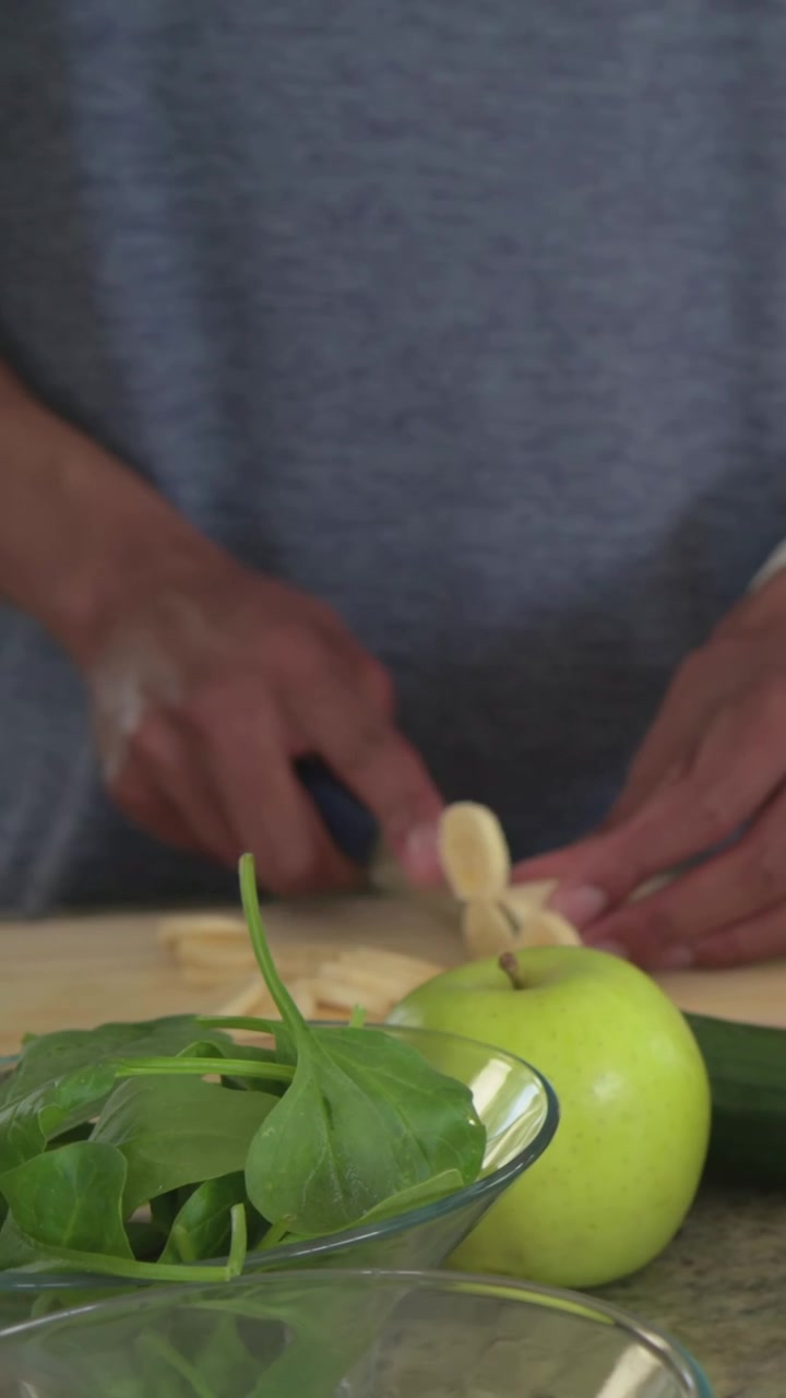 Video verticale: Uomo con maglietta grigia che tiene un coltello blu, affetta frutta su un tagliere, preparando un'insalata in cucina.