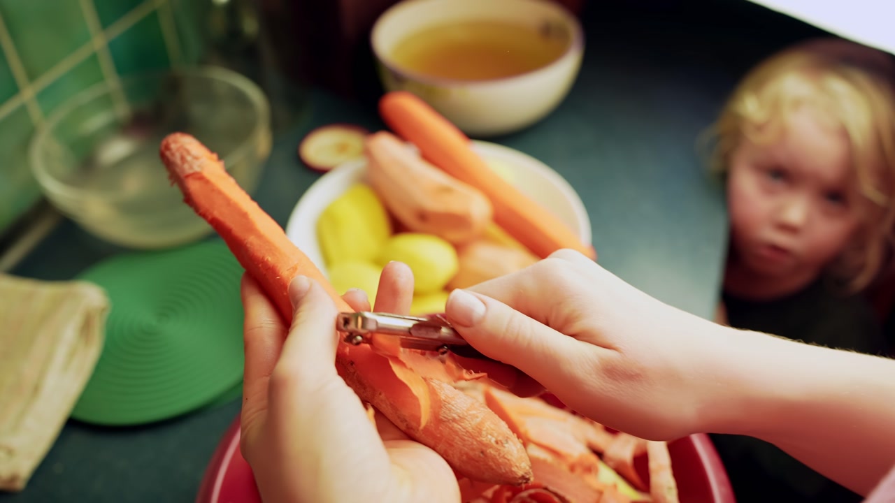 Close-up hands peeling a carrot in the kitchen while a toddler watches, everyday meal prep for a healthy family dinner and home lifestyle content.